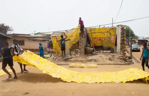 Nii Odzenma People spreading out yellow tapestry created by artist Serge Attukwei Clottey on a road in La - Accra, Ghana