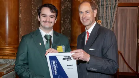 Harry Jones (elft) being handed an award by the Duke of Edinburgh, Harry is wearing a green suit, with a green tie and white shirt, he has short dark hair and a moustache, the Duke is wearing a grey pinstriped suit with a red tie, they are standing by pillars and curtains. Both men are smiling and looking straight at the camera. 