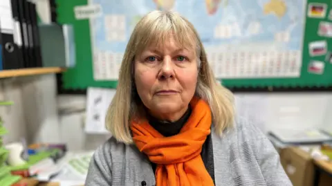 A woman with blonde shoulder length hair and fringe over her forehead is looking at the reader, wearing a grey cardigan and an orange scarf tied round her neck. She is sitting in her school classroom with a blurry image of a world map behind her and a desk with stationary on either side.