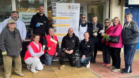 West Northamptonshire Council A large group of people all looking at the camera and smiling. Several a holding items like paint brushes, rollers, tools and plants. A sign is in the middle. Four people are kneeling down and the rest are standing up. Windows to a business are behind them. 