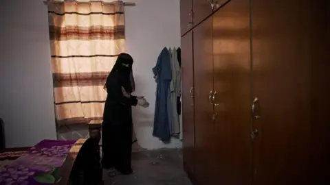 Safa wears a black headscarf, veil and robe covering everything except her hands and eyes. On the left is the foot of a double bed and on the right are some fitted wooden wardrobes. She points at the floor by the far end of the cupboards.