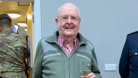 BBC Jack Waterfield smiling at the camera, with a glass of fizz in his hand. He has short white hair and glasses, and is wearing a green zip-up top over a white-and-red checked shirt. He is in a room with some people in military uniform around him. 