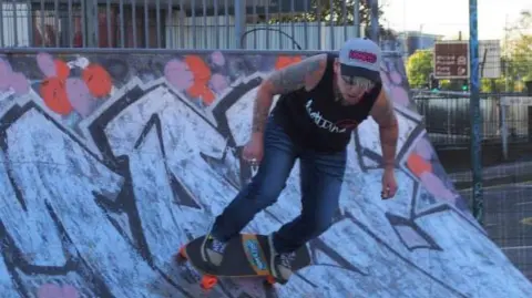 Skatenuts Ethel Keith Ewing rides a skateboard on a curved ramp at an outdoor skate park. She is wearing a dark sleeveless shirt with white lettering and blue jeans.