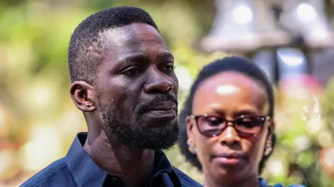 A close-up shot of Bobi Wine looking across the camera. He has a short beard and short hair. His wife is in the background, out of focus.