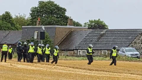 BBC Ten police officers wearing helmets standing in a field outside a row of properties. 