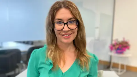 Rachel is sat in an office environment, and is smiling at the camera. She is wearing glasses, a green blouse and has long, wavy brown hair.
