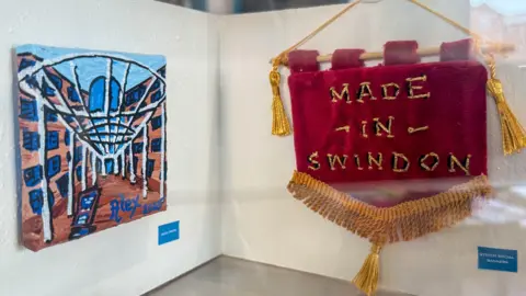 Tiny artwork on a tiny wall showing the local hospital beside a tiny embroidered banner saying 'made in swindon'.