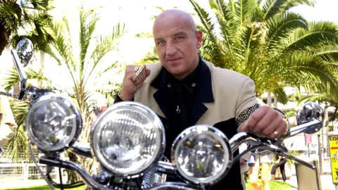 An image taken in Cannes in 2001 shows a younger Dave Courtney, a man with a bald head wearing a light jacket and black shirt, sitting on a motorbike with a close-up view of three round silver headlights on the handlebars. Small palm trees can be seen in the background