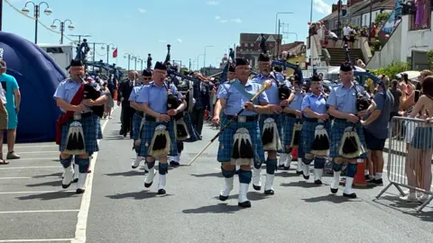 The Scottish pipers wear blue kilts and short-sleeved shirts with large tasseled sporrans. The leader carries a long ornamental pole at the front.