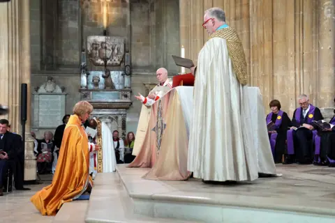 Stefan Rousseau / PA Media Mullally (left) kneeling before Dean of Canterbury, the Very Revd Dr David Monteith and Archbishop of York Stephen Cottrell all in robes while they install her as the new archbishop