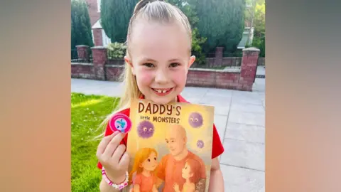 Isabella is holding her book and smiling at the camera. The front cover reads: Daddy's little monsters. There is a drawing of Isabella cuddling her dad Chris and her younger sister. There are three purple fluffy balls to represent the illness. 