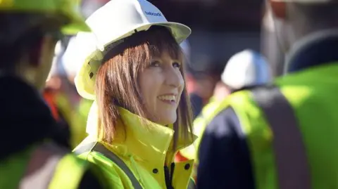 Rachel Reeves smiles wearing a white hard hat and a yellow fluorescent jacket. She has shoulder length brown hair with a fringe. She is speaking to men also in fluorescent jackets and white hard hats who are blurred in the foreground. 