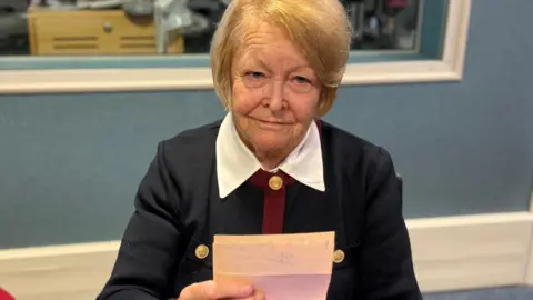 A picture of Penny holding a letter. She is wearing a black, red and white outfit and has blonde hair.