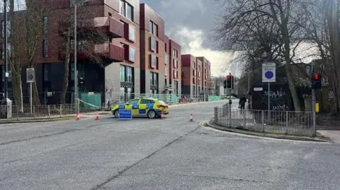 A shot of a street crossing, there is a police care across the road, as well as cones and police tape blocking the left exit off the road. To the far left there are buildings, with red brick at the top and black bricks at the bottom.