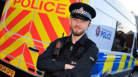 PC Doyle wearing a Greater Manchester Police has his arms folded and is smiling as he stands in front of the back of a white police van.