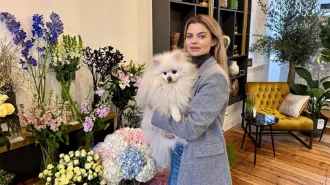 Alla Orekhovska inside her new flower shop holding a fluffy white dog. There are displays of a dozen different types of flowers behind her which are yellow, purple, pink and blue in colour. There is a chair and greenery in the background.