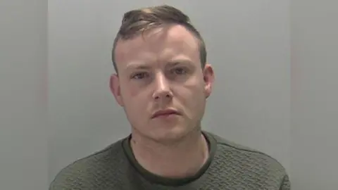 A mugshot of a man with short brown hair that is in a quiff. He is wearing a dark grey jumper and staring into the camera with a straight face. 
