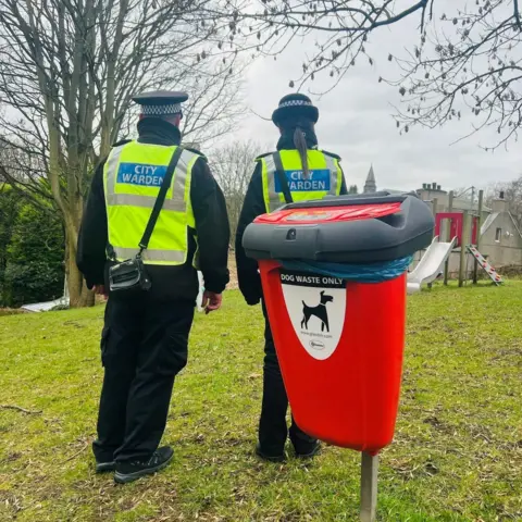 Two people with bright yellow 'City Warden' hi-vis jackets, standing on grass, behind a red dog waste bin.