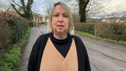 Nicky Brownjohn stood on Princes Road, Earlswood, Surrey. She has blonde hair and is wearing a black cardigan and tan top. 