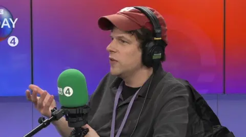 Jesse Eisenberg wearing red cap and headphones speaking into a mic in radio studio