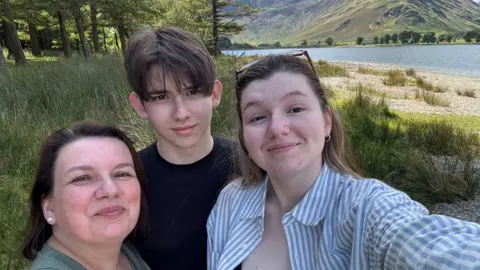 On the left Oles' mum Liudmyla, is smiling at the camera, she has short brown hair and brown eyes. Oles in the middle has on a black t-shirt and brown hair. On the right Oles sister Yaryna is wearing a white and blue striped shirt and has long light mousy brown hair. Behind them are huge mountains a lake and trees in the distance. 