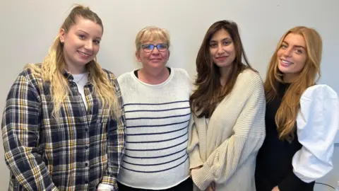 Shariqua Ahmed/BBC Nicole Dench-Layton, Heather Birch, Shamaila Mushtaq and Kayleigh Adams stand in a line smiling at the camera. Nicola is wearing a plaid shirt, Heather is wearing a striped jumper, Shamaila is wearing a beige long cardigan and Kayleigh is wearing a black pinafore dress with a white shirt.