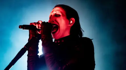 Marilyn Manson performs on stage, wearing black clothes and heavy make-up. His face, illuminated by a red spotlight against a blue stage background, is contorted while singing and shows metal teeth.