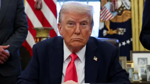 Reuters Donald Trump sitting at his desk wearing a dark suit and red tie.