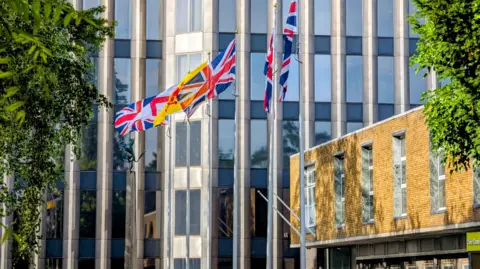 LDRS Several flags, including the Union Flag and a yellow flag featuring the Enfield Beast, fly on tall poles in front of the multi-storey glass and concrete facade of the Enfield Civic Centre. Part of a brick building and green trees frame the scene.
