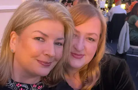 Family Two women next to each other are looking at the camera. People are at tables in the background.