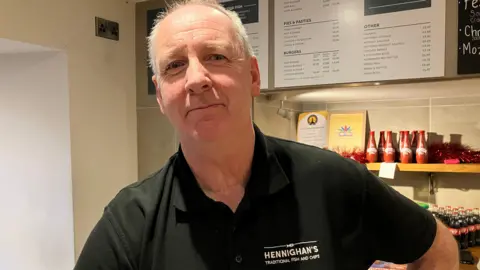 BBC A man is wearing a black polo and is working behind the counter of  fish and chip restaurant. Behind him are boards with costs, and various items such as bottles of vinegar.
