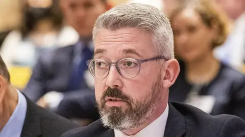 PA Niall Ó Donnghaile, pictured from the neck down, wearing a suit and white shirt. He has gray hair and wears glasses. 