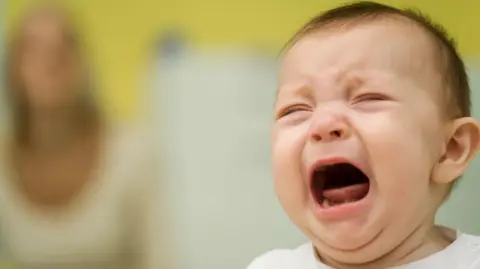 Baby crying with a mother out of focus in the background.