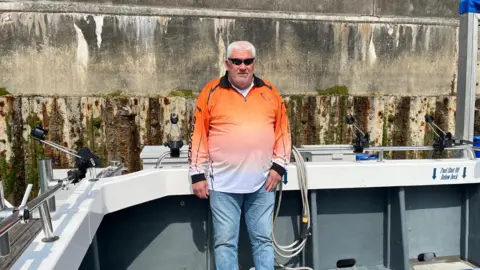 Joshua Askew/BBC A man with sunglasses and an orange top stands on a boat.