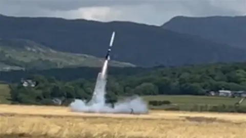 Rocket blasting into air with a cloud of smoke beneath it. In the background there are fields and green mountains.