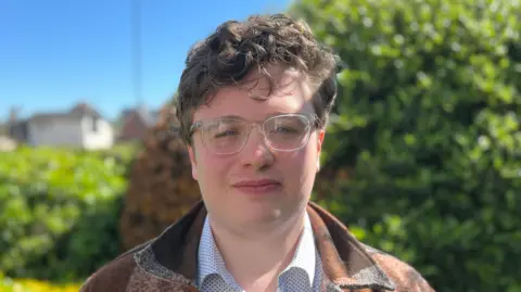Simon Dedman/BBC Kieron Franks stands outside on a sunny day in front of bushes. He has brown curly hair and wears glasses with clear frames, a brown jacket and polka dot shirt.