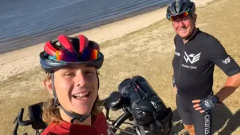 A selfie image of two men in cycling gear, one in red and one in black. They are standing by a beach