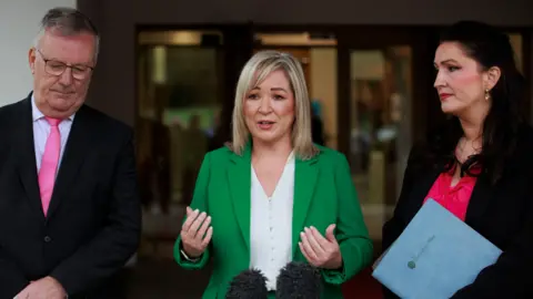 PA Media Mike Nesbitt, Michelle O'Neill and Emma Little-Pengelly standing around a microphone. Mike Nesbitt is wearing a black suit and pink tie with a white shirt. Michelle O'Neill is wearing a white shirt and green blazer while Emma Little-Pengelly is wearing a black blazer and pink top. She is carrying a blue folder.