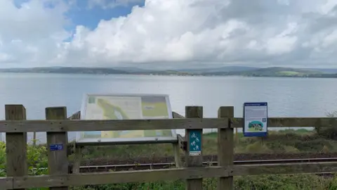 The University of Exeter A general view of a coastline behind a wooden fence. On the fence is a Voices of Exe Trail. It has writing on it and a piece of artwork and a QR code. Behind the fence is a sign about the Exe Estuary Trail. In the background is an estuary and hills.