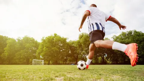 A man wearing football clothing running with a black and white football across an empty football field with a white goal in the background.