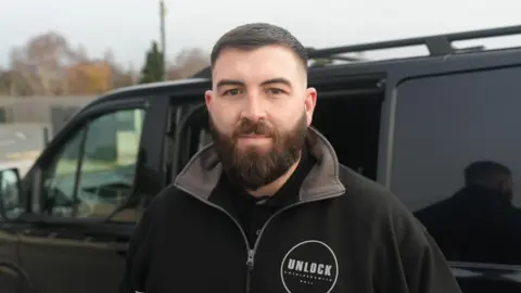 A man with dark hair and a beard looks straight to the camera. He is wearing a black jacket with the name of his company Unlock on the front