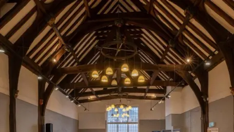 York Conservation Trust/City of York Council Interior of St Anthony's Hall. The ceiling is supported by thick wooden beams, with many branching up the walls. A large number of lights are hung from the ceiling beams, with a row of spotlights along each side. There is also a large window on the far wall.