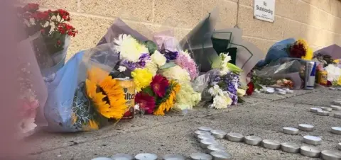 Bunches of flowers and candles are placed against a wall. Cans of drink are left too. Tealights spell out the names Mo and King with a heart depicted by tealights.
