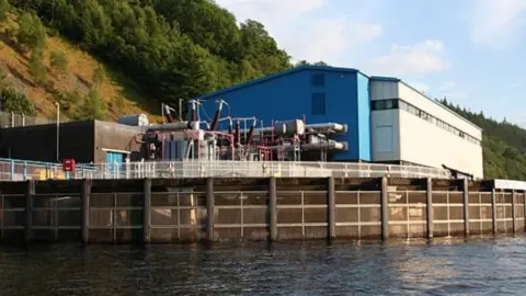 A large blue and white building with metal pipes and electrical cables coming out of it. The building is on the shore of the loch. There is a wooded hill behind it.  