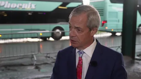 A man with grey hair, wearing a dark blue suit, white shirt and red tie. There are two green buses stationed behind him.