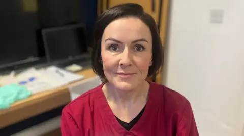 Lisa Ewart. She has short dark hair and brown eyes, wearing dark red scrubs.