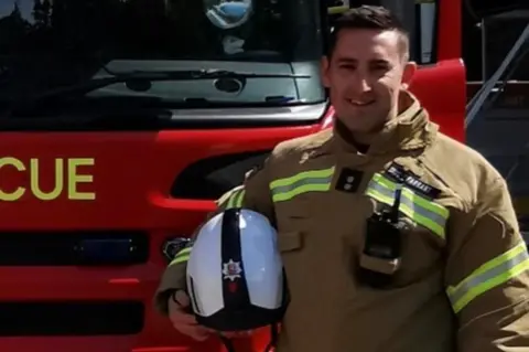 Essex County Fire and Rescue Service A man standing next to a fire engine with a suit on and a helmet under his arm
