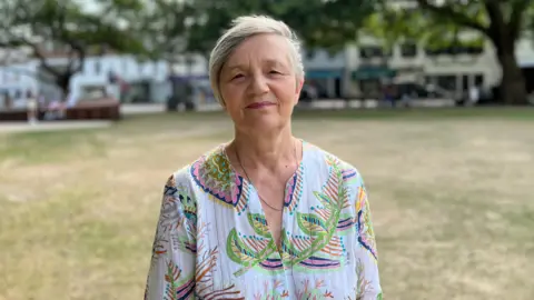 Mary Le Hegarat - a woman wearing a white long sleeve patterned blouse, standing in a public garden with blue skies.