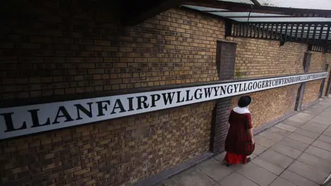 Getty Images Someone in Welsh costume on the platform of Llanfairpwllgwyngyllgogerychyndrobwllllantysiliogogogoch station, in front of the sign