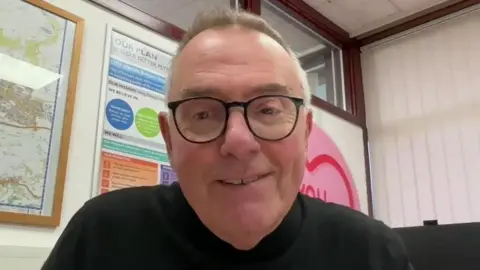 Plymouth City Council leader Tudor Evans smiles. He is wearing a black jumper and has black rimmed glasses on. He is sat in an office with posters and a map of Plymouth on the walls.